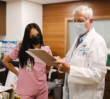 two masked medical professionals discussing looking at a clipboard and patient care